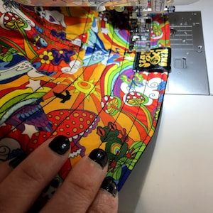 May include: A colorful fabric with a psychedelic pattern of mushrooms, rainbows, and flowers is being sewn on a sewing machine. The fabric has a black border and a black label with the text "Funky Socks" sewn on it.