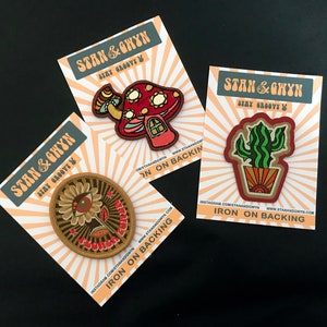 May include: Three iron-on patches with a retro, groovy aesthetic. The patches feature a mushroom house, a cactus in a pot, and a flower with an eye. Each patch has the text "Stan & Ghywn Stay Groovy" and instructions for ironing on the back.