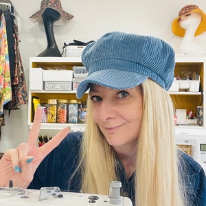 May include: A light blue corduroy newsboy cap with a short brim. The cap is worn by a person with long blonde hair. The background includes sewing supplies and other hats.