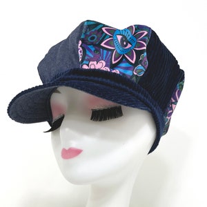 May include: A newsboy cap featuring a multi-panel design with dark blue denim, navy blue corduroy, and a vibrant floral print fabric in shades of purple, pink, and blue. The cap has a structured brim and a rounded crown, showcasing a mix of textures.
