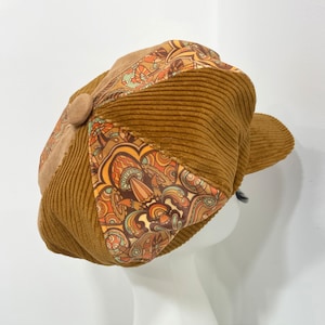 May include: A brown corduroy newsboy cap with a patterned fabric panel featuring a psychedelic design in shades of orange, yellow, and blue.