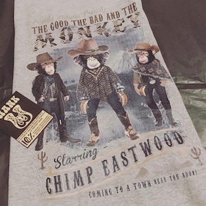 Può includere: Una maglietta grigia con una grafica di tre scimmie che indossano cappelli da cowboy e gilet. Il testo "Primate Pictures Presents The Good The Bad And The Monkey" è in cima alla grafica. Il testo "Starring Chimp Eastwood" è sotto la grafica. Il testo "Coming to a town near you soon!" è in fondo alla grafica.