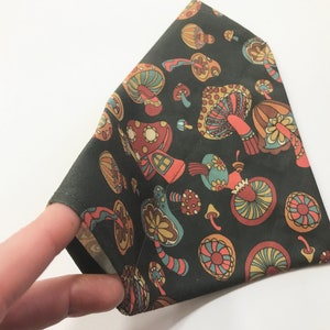 May include: A dark green fabric bandana with a mushroom print in shades of orange, brown, and blue. The mushrooms have a whimsical, retro design.