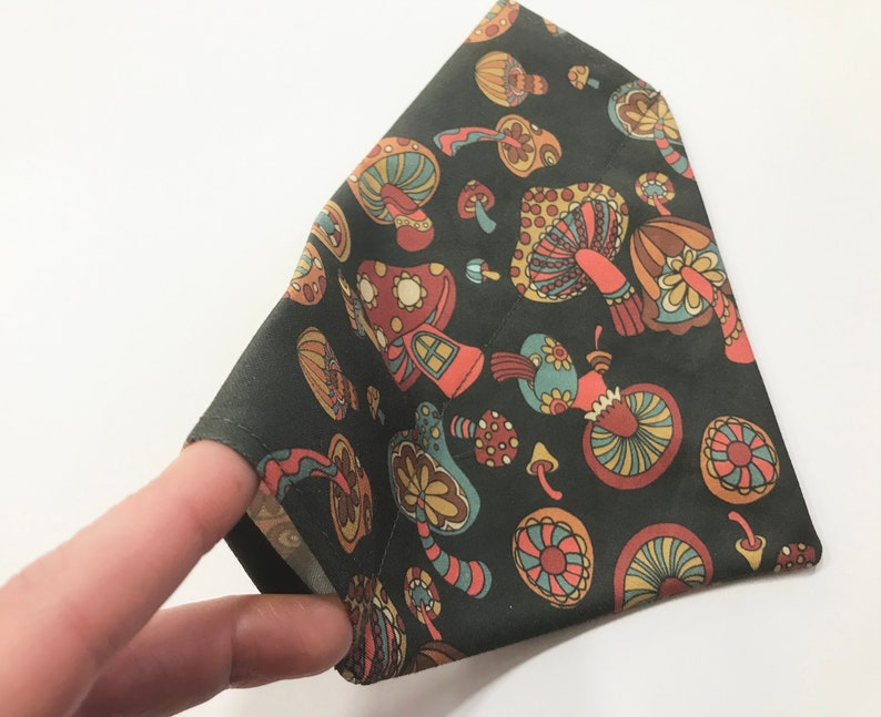 May include: A dark green fabric bandana with a mushroom print in shades of orange, brown, and blue. The mushrooms have a whimsical, retro design.