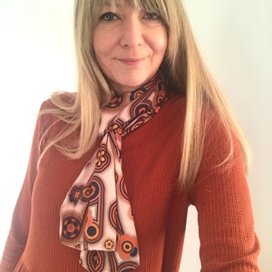 Pu&ograve; includere: Una donna indossa un abito a maniche lunghe color ruggine con una sciarpa a fantasia annodata al collo. La sciarpa ha un design geometrico con colori arancio, marrone e bianco.