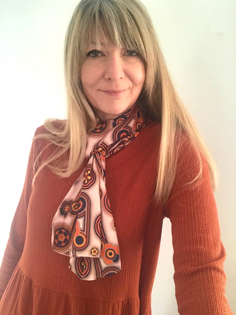 Pu&ograve; includere: Una donna indossa un abito a maniche lunghe color ruggine con una sciarpa a fantasia annodata al collo. La sciarpa ha un design geometrico con colori arancio, marrone e bianco.