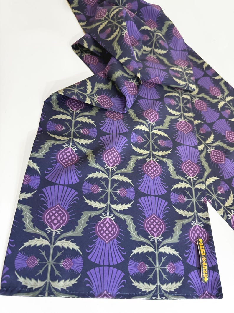 May include: A purple and green patterned silk scarf with a repeating thistle design. The scarf has a navy blue background and the text "OSCAR & ALEX" is printed on the bottom right corner.