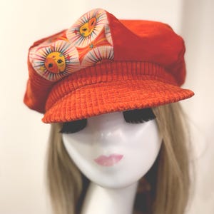 May include: Orange corduroy newsboy cap with a patterned sun design on the side. The cap has a short brim and is displayed on a mannequin head with blonde hair.