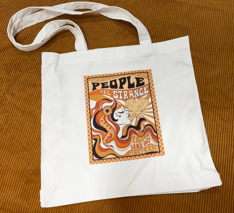 May include: White canvas tote bag featuring a retro-style graphic of a woman with long, wavy hair and the text "People are strange, Faces come out of the rain".
