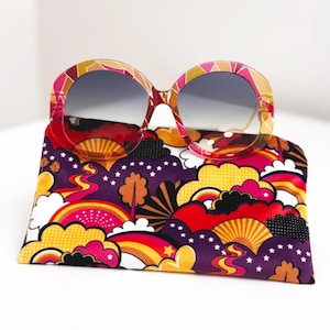May include: Oversized sunglasses with a patterned frame and grey lenses sit on a case. The case has a colourful design of rainbows, clouds, and stars in purple, red, yellow, and white. A retro style.