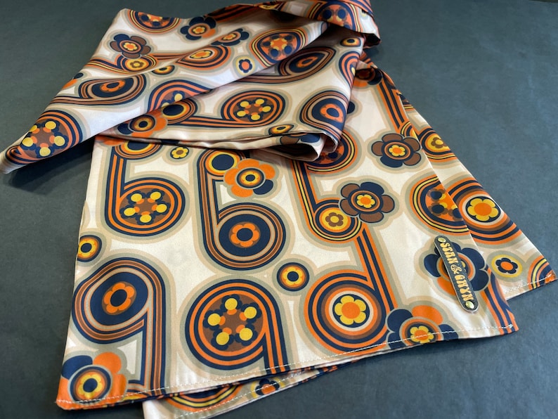 Pu&ograve; includere: Un foulard di seta beige con un motivo retr&ograve; di cerchi e fiori arancioni, marroni e blu. Il foulard ha un piccolo cartellino dorato con il testo "seam & onyx".