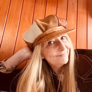 May include: A multi-toned leather newsboy cap with a quilted brim and a cream-coloured side panel. The cap has a brown leather top and a dark brown leather accent. The hat is a stylish accessory.