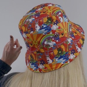 May include: A colorful bucket hat with a psychedelic pattern featuring mushrooms, rainbows, and cartoon animals. The hat is orange, red, yellow, blue, green, and white.