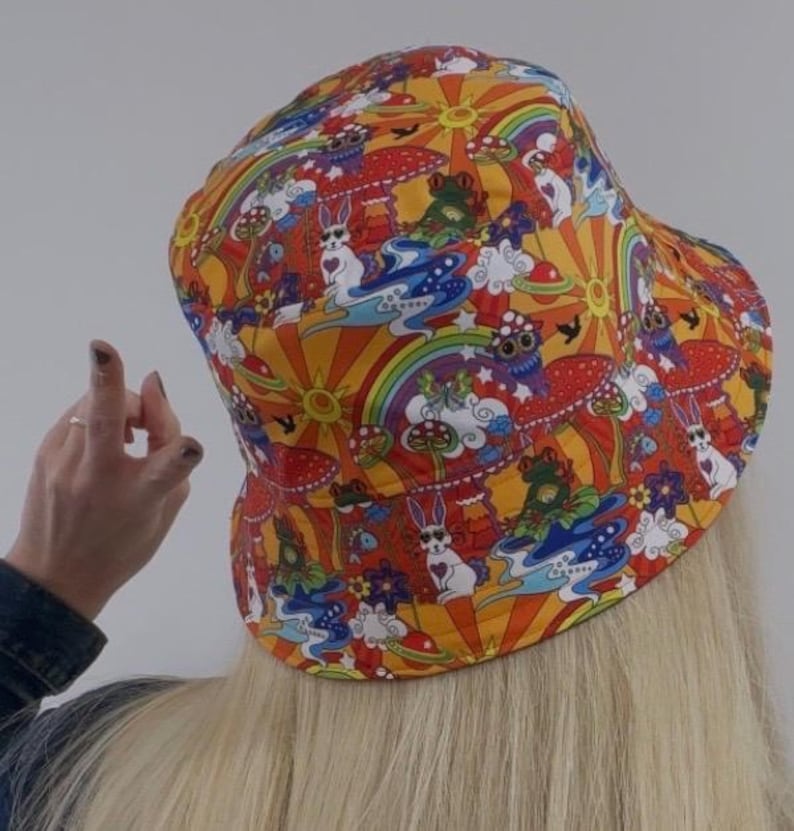 May include: A colorful bucket hat with a psychedelic pattern featuring mushrooms, rainbows, and cartoon animals. The hat is orange, red, yellow, blue, green, and white.