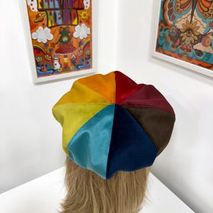 May include: A multi-colored beret constructed from eight triangular panels of velvet fabric. The hat features a vibrant spectrum of colors including orange, yellow, lime green, light blue, dark blue, brown, maroon, and red, arranged in a circular pattern.
