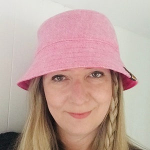 May include: A woman wearing a pink bucket hat with a braided hairstyle. The hat is made of a soft, fuzzy material.