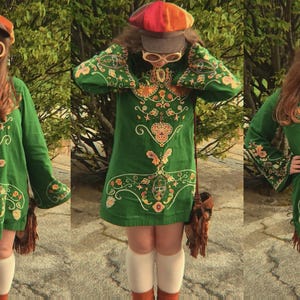 May include: A green long-sleeve tunic dress featuring intricate gold and orange embroidery, paired with a multi-colored newsboy cap, large round cream-colored sunglasses, white knee-high socks, and a brown fringed shoulder bag.