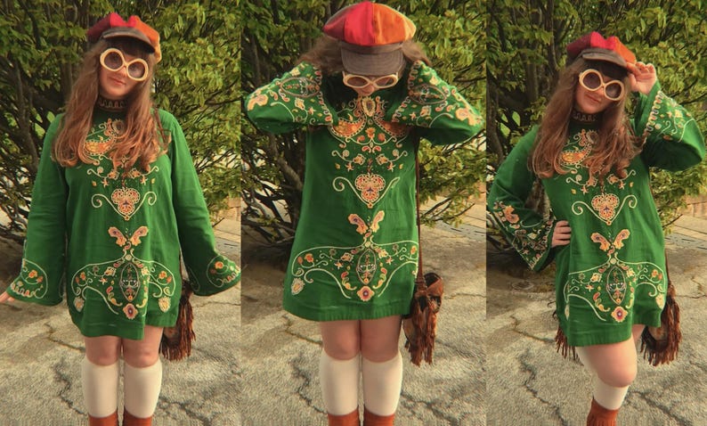 May include: A green long-sleeve tunic dress featuring intricate gold and orange embroidery, paired with a multi-colored newsboy cap, large round cream-colored sunglasses, white knee-high socks, and a brown fringed shoulder bag.
