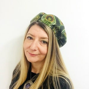 May include: A woman with long blonde hair is wearing a green and brown patterned beret. She is wearing a black shirt and a silver necklace with a large purple stone pendant.