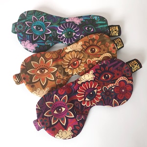 Könnte beinhalten: Drei Augen-Themen-Schlafmasken mit Blumenmustern. Die Masken sind aus Stoff und haben ein dunkles Grün-, Braun- und Violett-Farbschema. Jede Maske hat ein goldenes Etikett mit dem Text "Stay & Dream".