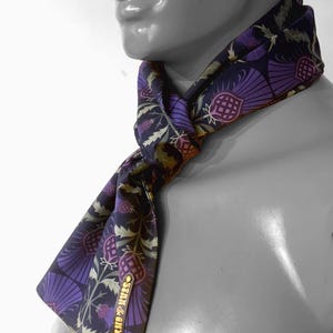 May include: A purple and green patterned silk scarf with a gold label that reads "STAIN & GREY" tied around a mannequin's neck.