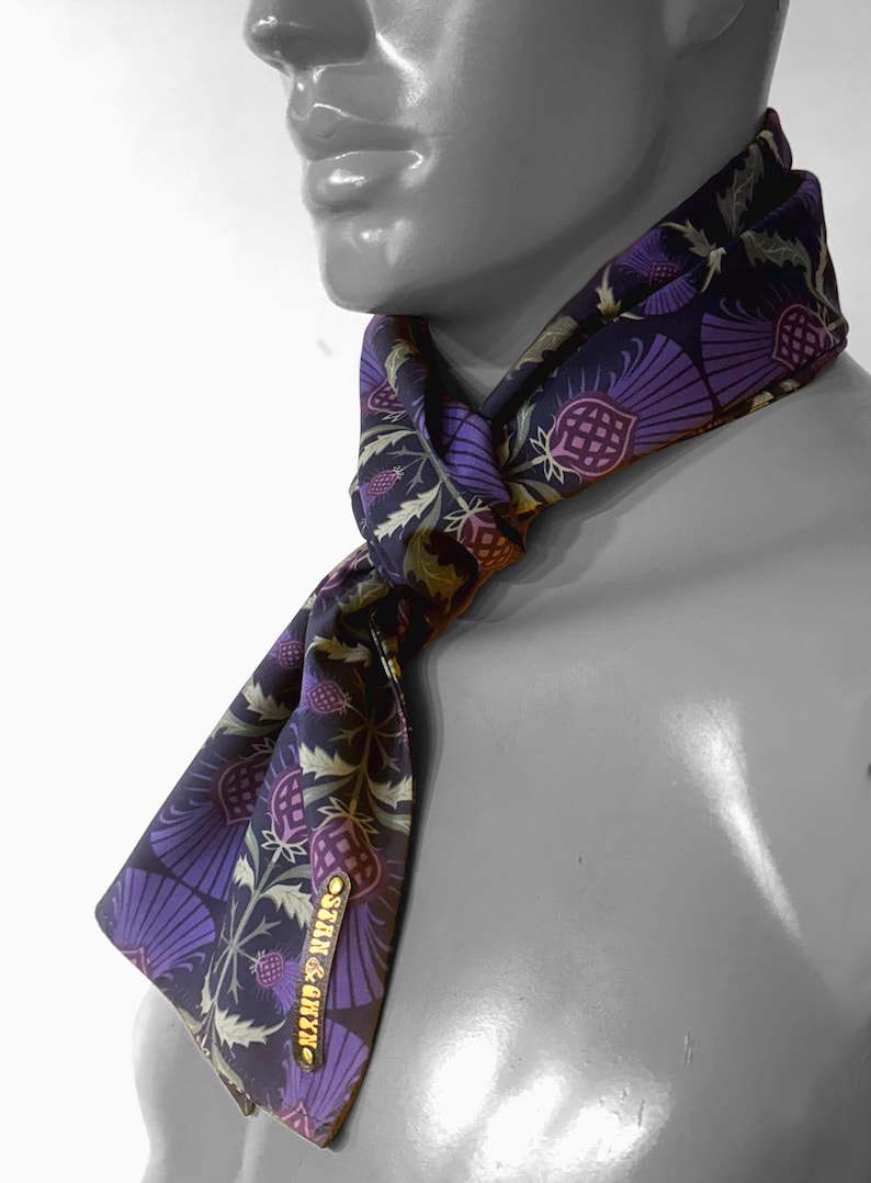 May include: A purple and green patterned silk scarf with a gold label that reads "STAIN & GREY" tied around a mannequin's neck.
