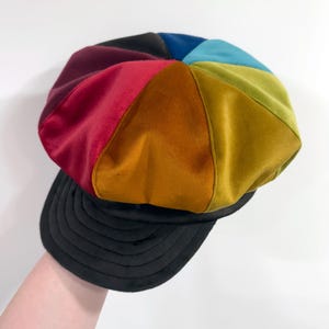 May include: A multi-colored newsboy cap featuring panels of velvet fabric in shades of deep red, orange, mustard yellow, olive green, teal, and black. The hat has a rounded crown constructed from eight triangular segments and a black brim.