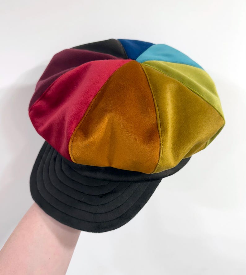 May include: A multi-colored newsboy cap featuring panels of velvet fabric in shades of deep red, orange, mustard yellow, olive green, teal, and black. The hat has a rounded crown constructed from eight triangular segments and a black brim.