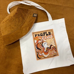May include: A white tote bag with a brown corduroy bucket hat. The tote bag features a graphic design with the text "People are strange" and a woman with a flower in her hair. The text "Rains come out of the rain" is also on the bag.