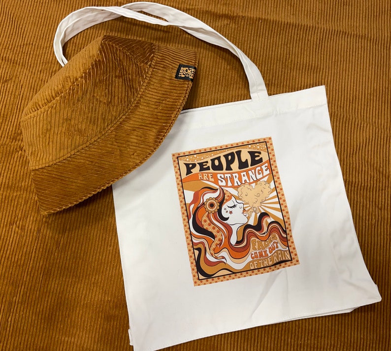 May include: A white tote bag with a brown corduroy bucket hat. The tote bag features a graphic design with the text "People are strange" and a woman with a flower in her hair. The text "Rains come out of the rain" is also on the bag.