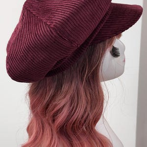 May include: A deep burgundy corduroy newsboy cap featuring a structured crown and a short, stiff brim. The fabric has a distinct vertical ribbed texture throughout the entire surface of the hat, providing a consistent look across the top and the visor.