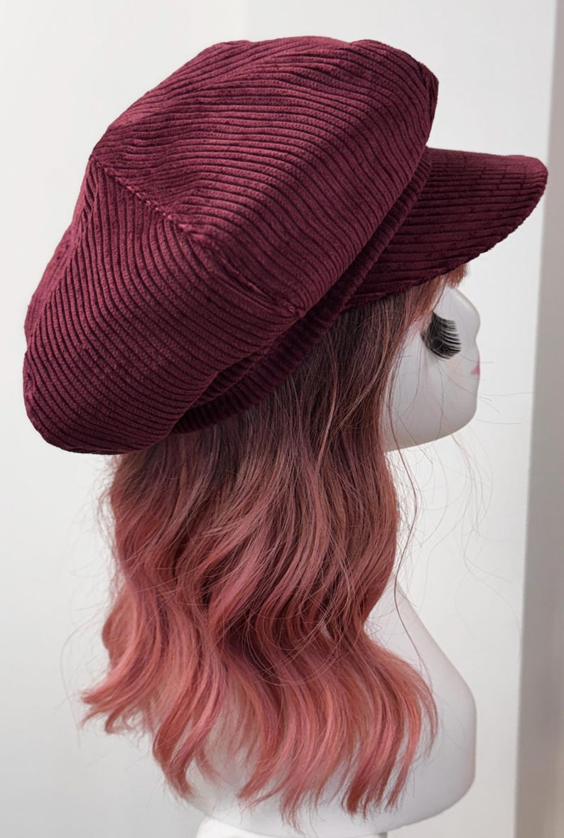 May include: A deep burgundy corduroy newsboy cap featuring a structured crown and a short, stiff brim. The fabric has a distinct vertical ribbed texture throughout the entire surface of the hat, providing a consistent look across the top and the visor.