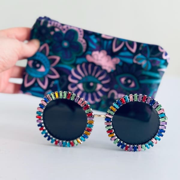 May include: Round sunglasses with black lenses and a gold frame. The rims are adorned with colorful rectangular gemstones. A patterned pouch with blue, pink, and teal floral designs is in the background.