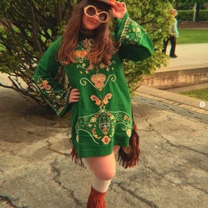 May include: A green long-sleeved tunic dress featuring intricate multi-colored embroidery, paired with white tights, brown fringed ankle boots, a multi-colored cap, and large round sunglasses with light-colored frames and dark lenses.