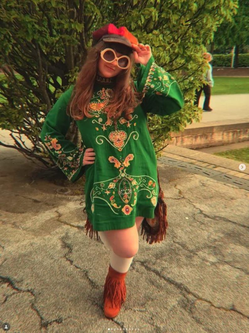 May include: A green long-sleeved tunic dress featuring intricate multi-colored embroidery, paired with white tights, brown fringed ankle boots, a multi-colored cap, and large round sunglasses with light-colored frames and dark lenses.