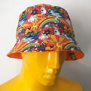 May include: A bright orange bucket hat with a psychedelic print featuring rainbows, stars, clouds, and cartoon animals. The hat has a black and white label with the text "Vegan & Cruelty Free".