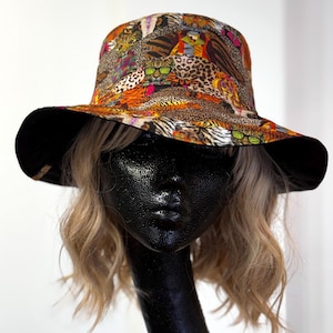 May include: A patterned bucket hat with a black brim. The hat features a colourful animal print design, including tigers and leopards. The hat is displayed on a black mannequin head, with blonde hair.