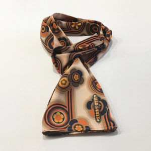 Pu&ograve; includere: Un foulard di seta beige con un motivo floreale e geometrico retr&ograve; in tonalit&agrave; arancio, marrone, giallo e nero. Il foulard ha un'etichetta dorata con il testo "STAN & CHYZ".