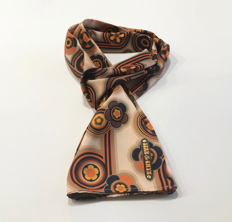 Pu&ograve; includere: Un foulard di seta beige con un motivo floreale e geometrico retr&ograve; in tonalit&agrave; arancio, marrone, giallo e nero. Il foulard ha un'etichetta dorata con il testo "STAN & CHYZ".