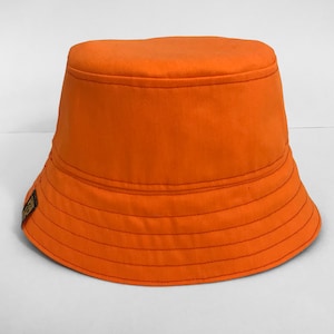 May include: An orange bucket hat with a slightly curved brim. The hat has a single stitch detail around the brim and a small brown patch on the side.