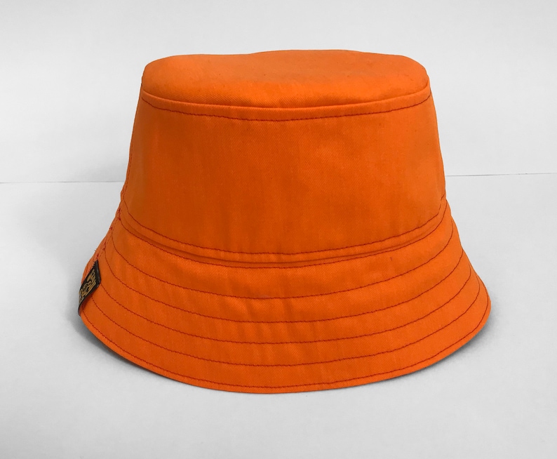 May include: An orange bucket hat with a slightly curved brim. The hat has a single stitch detail around the brim and a small brown patch on the side.