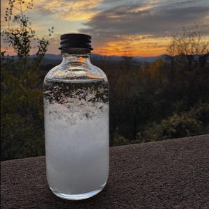 May include: A clear glass bottle with a black cap, filled with a cloudy, white substance. The bottle sits on a brown surface, with a scenic sunset in the background. The sky is filled with orange, yellow, and blue hues, and the trees are silhouetted.