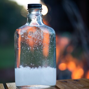May include: A clear glass bottle with a black cap, filled with a cloudy liquid. The bottle sits on a wooden surface, with a blurred background of green and orange hues. The liquid appears to have a white sediment at the bottom.