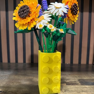 May include: A bright yellow vase made of interlocking blocks holds a colorful bouquet of flowers. The flowers include sunflowers, daisies, and lavender. The sunflowers have yellow petals and brown centers. The vase is rectangular with raised circular studs.