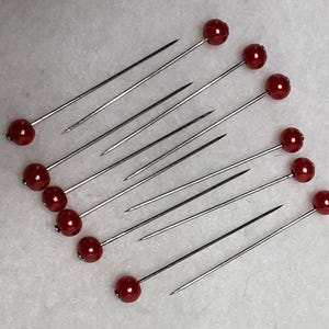 May include: A collection of sewing pins with long, silver shafts and round, red, bead-like heads. The pins are arranged diagonally across a light-colored surface, showcasing their sharp points and decorative tops. These pins are likely used for crafting or sewing projects.