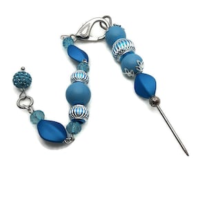 May include: A blue beaded necklace with a silver clasp and a silver pin. The necklace has a variety of blue beads, including round, oval, and faceted beads. The pin is attached to the necklace with a silver loop.