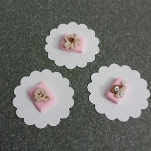 May include: Three white scalloped circles with pink squares and brown fondant decorations. The decorations are different on each square.