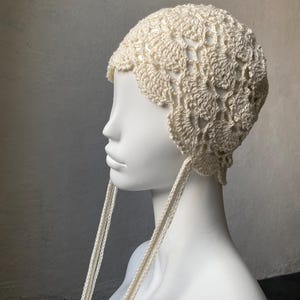 Pode incluir: Chapéu de crochê marfim com borda ondulada e longas amarrações trançadas. O chapéu apresenta um design aberto e rendado. O chapéu é exibido em uma cabeça de manequim branca.