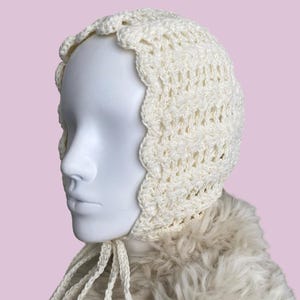 May include: A white crocheted baby bonnet with a scalloped edge and a tie. The bonnet is on a white fur-like surface.