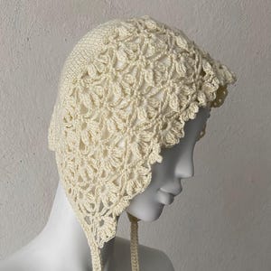 May include: Cream-colored crochet bonnet with a floral pattern. The bonnet features a scalloped edge and ties under the chin. The handmade hat is displayed on a mannequin head against a neutral background.